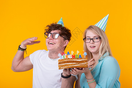 有趣的年轻夫妇戴着纸帽 拿着蛋糕 站在黄色背景下 做着傻脸 祝生日快乐 祝贺和鬼混的概念风格蓝色派对惊喜情绪潮人创造力喜悦工作室背景图片