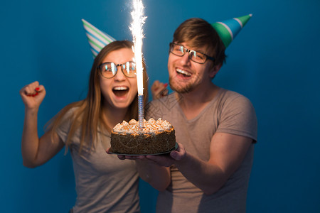 有趣的年轻夫妇戴着纸帽 拿着蛋糕 站在蓝色背景下 做出一张傻脸 祝生日快乐 祝贺和鬼混的概念甜点工作室周年乐趣喜悦礼物娱乐风格庆背景图片