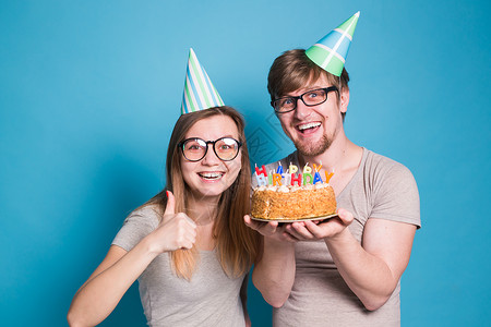 有趣的年轻夫妇戴着纸帽 拿着蛋糕 站在蓝色背景下 做出一张傻脸 祝生日快乐 祝贺和鬼混的概念风格潮人装饰惊喜艺术喜悦节日工作室周背景图片