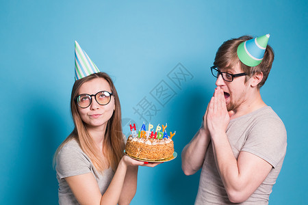 有趣的年轻夫妇戴着纸帽 拿着蛋糕 站在蓝色背景下 做出一张傻脸 祝生日快乐 祝贺和鬼混的概念礼物情绪生日喜悦甜点乐趣周年海报装饰背景图片