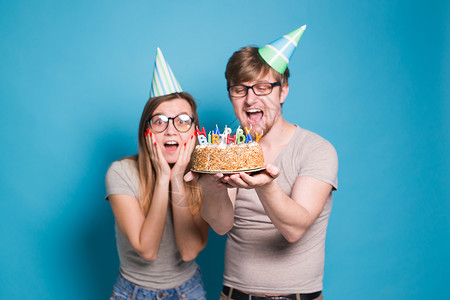 有趣的年轻夫妇戴着纸帽 拿着蛋糕 站在蓝色背景下 做出一张傻脸 祝生日快乐 祝贺和鬼混的概念节日风格乐趣礼物情绪惊喜娱乐喜悦庆典背景图片