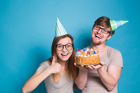 有趣的年轻夫妇戴着纸帽 拿着蛋糕 站在蓝色背景下 做出一张傻脸 祝生日快乐 祝贺和鬼混的概念生日纪念日情绪海报喜悦创造力艺术娱乐背景图片