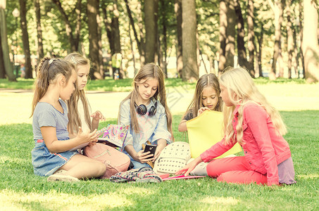 坐在公园里友善的青少年阅读女朋友手机喜悦朋友们童年友谊幸福草地阳光背景图片
