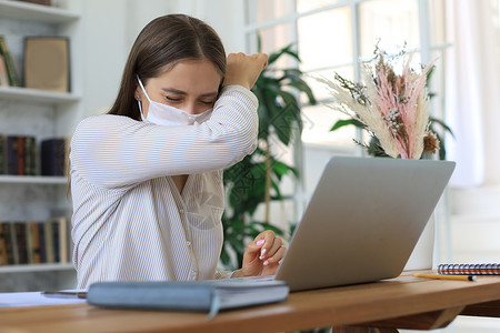 戴医疗保护面罩的女青年在自孤立和隔离期间在家里用笔记本电脑工作 笑声流感症状工人感染药品女士办公室疾病笔记商业在家办公高清图片素材