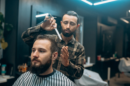 快乐的留着胡子的年轻男子在理发店被理发师理发男性沙龙梳子胡须治疗发型工作顾客服务店铺剪刀高清图片素材