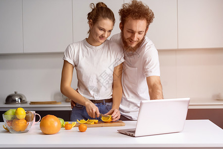 男人靠在女孩身上微笑着看浪漫电影 美丽的年轻夫妇使用笔记本电脑进行视频通话 年轻夫妇在家里的厨房里做健康的食物幸福丈夫桌子男性成男朋友高清图片素材