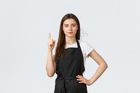 杂货店员工 小型企业和咖啡店的概念 穿着黑色围裙的年轻可爱女咖啡师手指向上展示广告 售货员提供资料购物高清图片素材