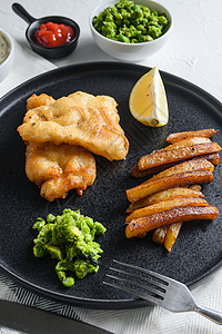 英国传统炸鱼薯条配薄荷豌豆泥和一片柠檬 在白色棉布和石头表面的黑色圆盘上英国食品高清图片素材