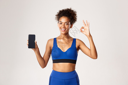 运动和技术概念 满意的非洲-美国女运动员穿蓝色健身服 显示智能手机屏幕和好手牌 白底白底等信息背景图片