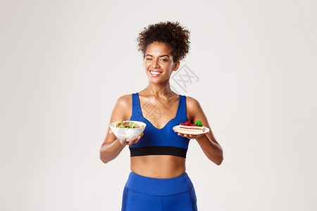 健康生活方式和体育理念 蓝色运动中的快乐女运动员 展示了沙拉和蛋糕的盘子 站在白背景上 笑声皮肤身体活力健身房衣服锻炼动机女性女背景图片