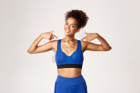 健康而健康的非洲裔美国健身女孩的腰部 穿着运动服 带着高兴的微笑指着自己 展示锻炼进展 白色背景背景图片