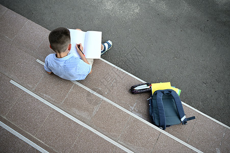 小学生专注于做家庭作业 坐在学校大楼的楼梯上 旁边是他的铅笔盒 里面放着学习用品 背包里放着工作簿 回到学校的概念背景图片