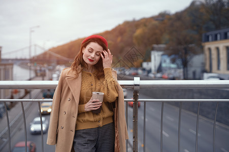 城市街道上拿着咖啡杯的女人 迷人的女人享受她的咖啡站在一座春天的城市桥梁与后面的交通 红色贝雷帽的法国妇女有都市背景时尚成人技术背景图片