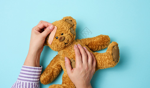 动物援助雌性手握着棕色泰迪熊 在蓝色背景上粘贴医学粘合石膏 电车治疗疾病治愈娃娃创伤动物儿科医院援助伤害疼痛背景