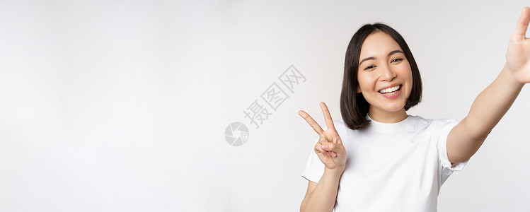 美丽的亚洲年轻女性自拍 用和平 V 字牌摆姿势 开心地笑着 拍照 在白色背景下摆姿势工作室大学互联网学生情绪女士横幅情感技术女朋背景