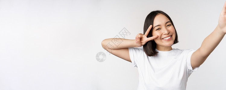美丽的亚洲年轻女性自拍 用和平 V 字牌摆姿势 开心地笑着 拍照 在白色背景下摆姿势衬衫工作室技术学生女士女朋友企业家情绪成人大背景