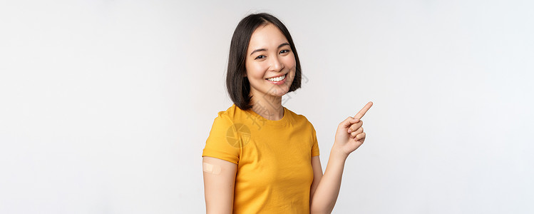Covid19 和疫苗接种概念 微笑的亚洲女孩戴着肩带 用手指指着横幅 展示疫苗运动 站在白色背景上购物女朋友互联网女士企业家情背景图片