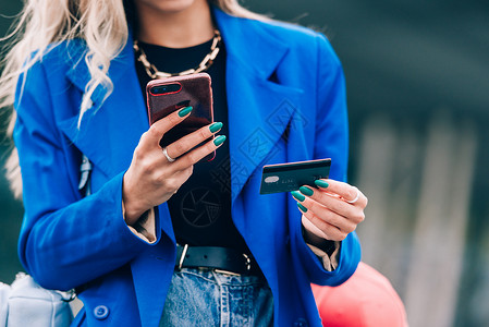 穿着蓝色夹克的时装金发美女小飞人 拿着信用卡和智能电话在街上买单工具卡片成人技术女士金发女郎手机银行购物女孩背景图片
