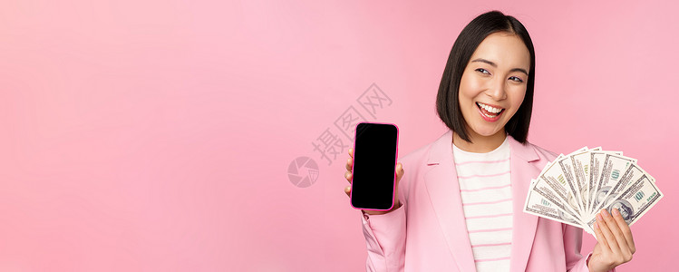 小额信贷字体成功的年轻亚洲女商务人士展示金钱 现金和智能手机屏幕 微笑着高兴 粉红色背景女士银行业银行标识商务文员信用投资细胞工作室背景