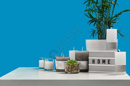 白色的桌子上放着绿色植物和多汁的盆栽 一些不同大小和颜色的蜡烛 灯上刻着家的字样 蓝色背景 特写背景图片