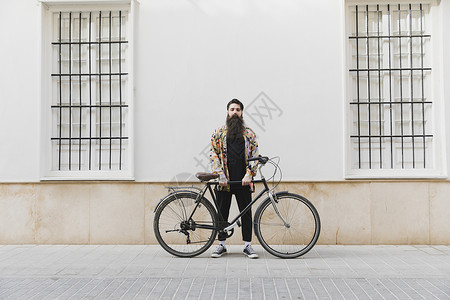 带着自行车靠墙站着的长胡子的青年男子牛仔裤高清图片素材