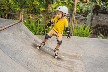拿滑板男孩戴头盔和膝盖垫的体育男孩学会在滑板公园与滑板一起玩滑板城市游戏运动鞋溜冰者街道学习行动太阳闲暇青少年背景