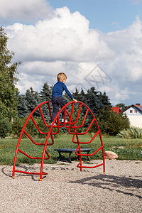 一个非常勇敢的小男孩在操场上爬上一个红色的金属大拱形梯子 儿童游戏中父母关爱和安全的概念孩子平衡假期运动时间训练活动儿子学习晴天背景图片