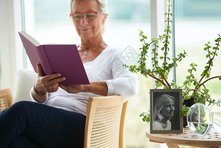 一位在休息室读一本好书的年长女子 和一张她自己的旧相片一起阅读背景图片