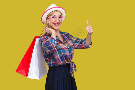 时尚的中年妇女 在黄色背景下从事临时作风购物祖母店铺销售乐趣衬衫拇指手指幸福老年背景图片