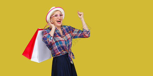 时尚的中年妇女 在黄色背景下从事临时作风衬衫成功老年祖母成人购物幸福顾客帽子尖叫背景图片