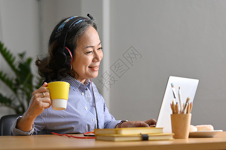 持着咖啡杯和观看笔记本电脑网上网络研讨会的耳机微笑成年妇女搜寻高清图片素材