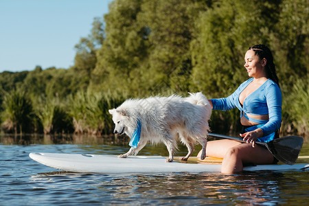 划船的女孩年轻女子与她的宠物一起在湖上划船 雪白狗养小日本人Spitz站在Sup委员会中晚饭女孩成年木板池塘城市动物蓝色女性游泳背景