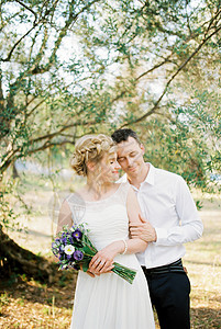 用橄榄树附近的花束从后面抱着新娘的Groom拥抱背景图片