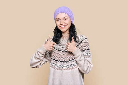 穿着冬季毛衣帽的快乐年轻女子 在棕色背景上举起拇指帽子高清图片素材