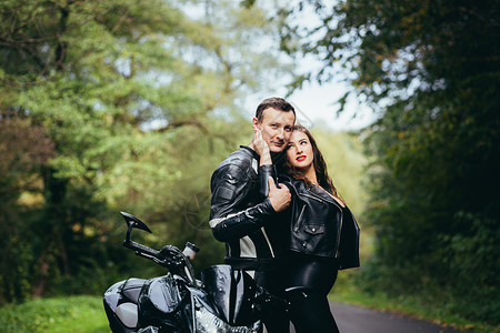 英俊的年轻男子和美丽的年轻女孩 一对夫妇坐在摩托车上 站在一辆穿着黑色皮衣的摩托车旁边 在大自然中拥抱 在街上男人假期运输女朋友男朋友高清图片素材