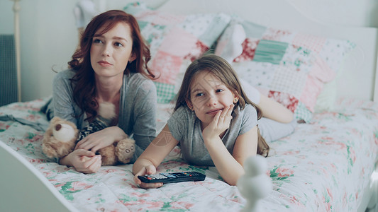 卡通女孩和熊快乐的母亲和可爱的女儿在早上躺在家里舒适的卧室里 用遥控器和微笑在电视上看卡通电影背景