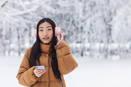 粉色电话美丽的日本青少年在冬季公园散步时使用电话 并用可爱的粉红耳机听音乐和感动的粉红色耳机女士雪花耳朵学生音乐技术手机微笑歌曲粉色背景