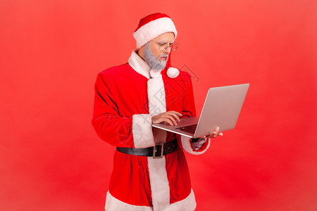 有灰胡子的老年男子 穿着圣达克萨斯服装 在电脑上工作 看着显示 集中的面部表情表现背景图片