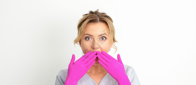 一位年轻的天主教女医生或护士的肖像被用粉色手套手在白色背景上蒙住嘴而震惊 笑声诊所金发女郎职业药剂卫生手势药品保健女性外套健康高清图片素材