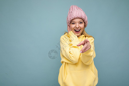 我粉色帽子照片中 美丽 微笑的年轻金发女子被隔离在蓝色背景墙上 戴着冬季粉色帽子和黄色连帽衫 看着相机 指着你背景