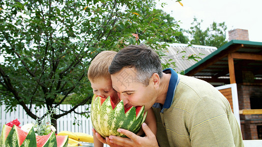 坐着西瓜的孩子夏天 在花园里 父亲带着四岁的儿子切西瓜吃 玩得很开心 一个男孩非常喜欢西瓜 与家人共进午餐的甜西瓜孩子女性营养白色姐姐动作快乐背景