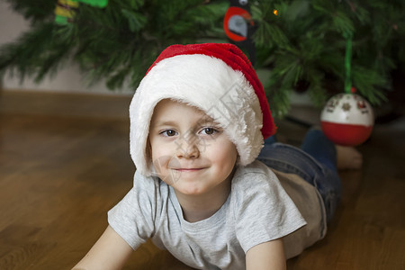 圣诞节舞台一张穿着灰色 T 恤和圣诞老人帽子的漂亮男孩在圣诞树上看着镜头的照片 在明亮的房间里的肖像 自然的 而不是舞台摄影玩具幸福喜悦年背景