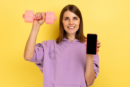 女性持有空屏幕智能手机和粉红色哑铃 广告宣传新的健身应用软件背景图片