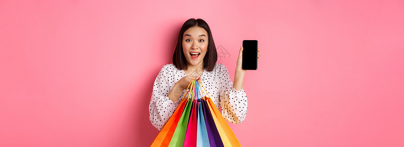 迷人的亚洲女性展示智能手机应用程序和购物袋 通过应用程序在线购买 站在粉红色背景中屏幕网络电话黑发互联网女孩广告店铺学生技术背景图片