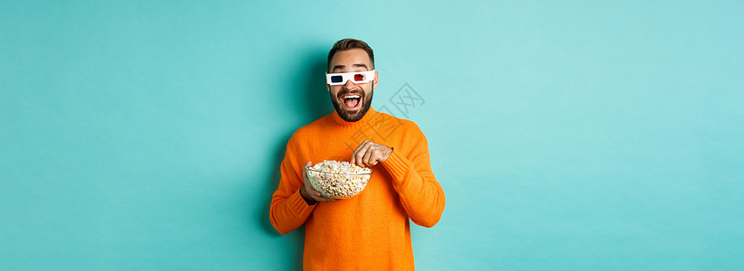 喜剧电影 看电视屏幕和吃爆米花 站在蓝背景上站立着的3D眼镜欢乐和惊异的年轻人横幅男性潮人广告橙子快乐黑发男人标识情感背景图片