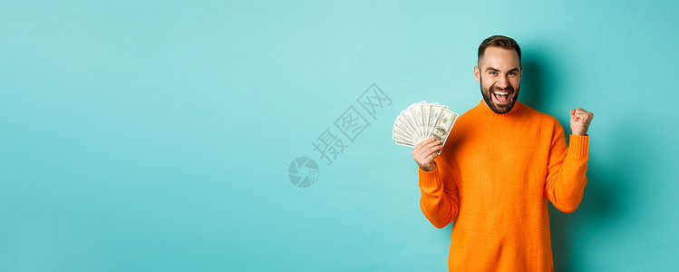 欢乐的人拿着钱 赢得现金奖和拳击泵 以满意的表情取胜 站在橙色毛衣与绿绿背景对立喜悦快乐横幅手势黑发工作室男人胡子橙子现金背景图片