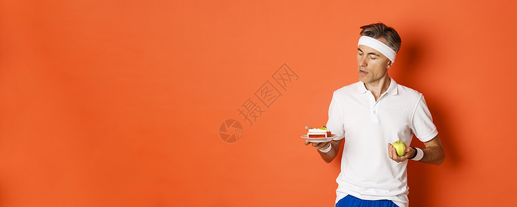 中年运动运动员在绿苹果和蛋糕之间做决定的肖像 正在吃饮食 站在橙色背景之上促销工作室胡须男性橙子健身房成人广告饮食训练背景图片