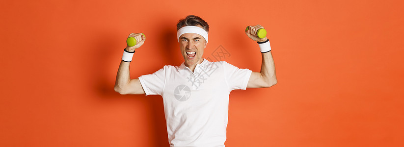 充满自信的中年健身人肖像 在橙色背景 柔力二头肌和握着哑铃进行体育锻炼训练广告男性运动服灰色工作室情感运动员促销橙子背景图片