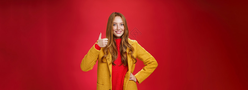 高兴和满意的红发女顾客竖起大拇指表示赞同和认可 对站在红色背景上的新黄色外套的优质质量感到满意 笑得非常开心自信成人理发新年快乐背景图片