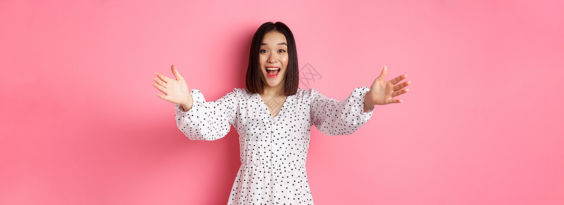 快乐的亚洲女孩伸手对着镜头 伸手去拥抱 微笑着想拿东西 站在粉色背景的裙子上黑发广告促销工作室青少年女性购物学生情绪冒充背景图片